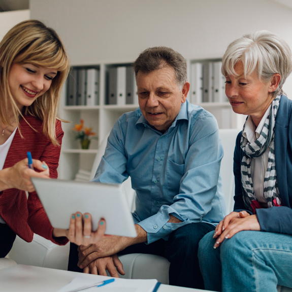 Photo of a woman holding a tablet with a seated man and woman looking on  the tablet
